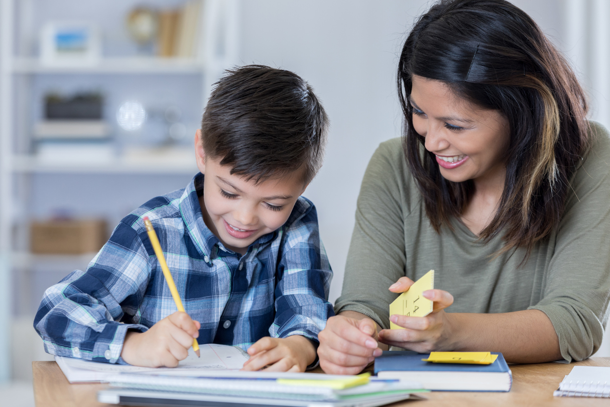 Cheerful mid adult Asian mom teaches her son math at home. The boys is writing something in a workbook. His mom is holding math flashcards.